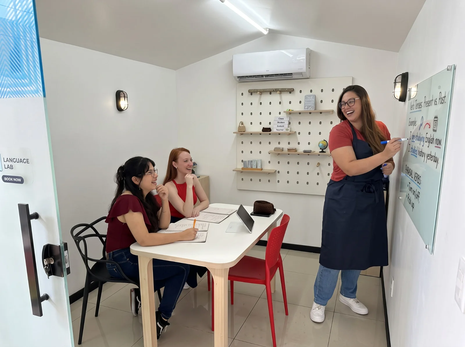 Duas alunas em aula em dupla na sala Language Lab da Tell Me, com a professora escrevendo no quadro branco