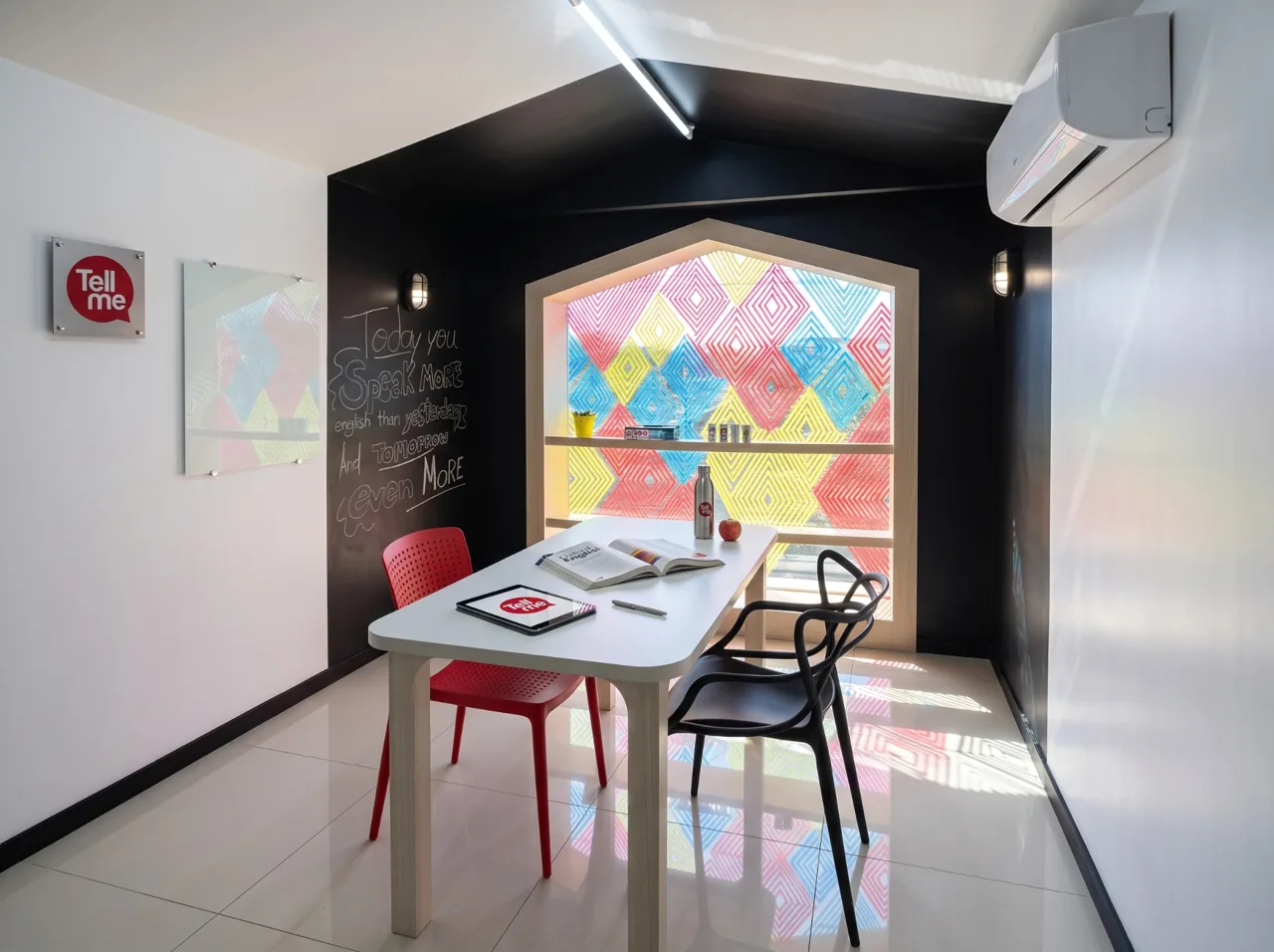 Sala de aula da Tell Me em Cascavel com mesa branca, cadeiras vermelha e preta, parede preta com lousa e vitral colorido da marca