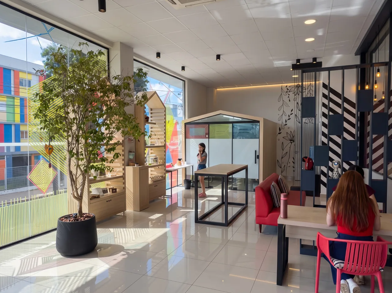 Hall de entrada da Tell Me em Cascavel com árvore em vaso, estante de madeira, mesas altas e poltrona vermelha junto às janelas de vidro
