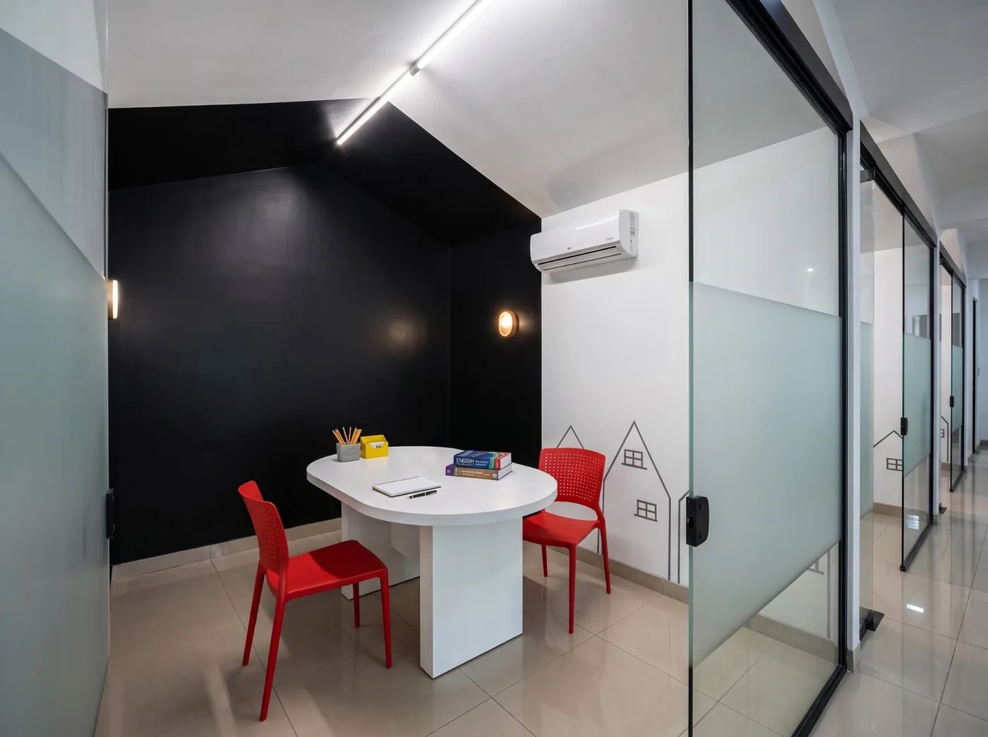 Sala de aula da Tell Me em Toledo com mesa branca oval, cadeiras vermelhas, livros e parede preta, ao lado do corredor de vidro