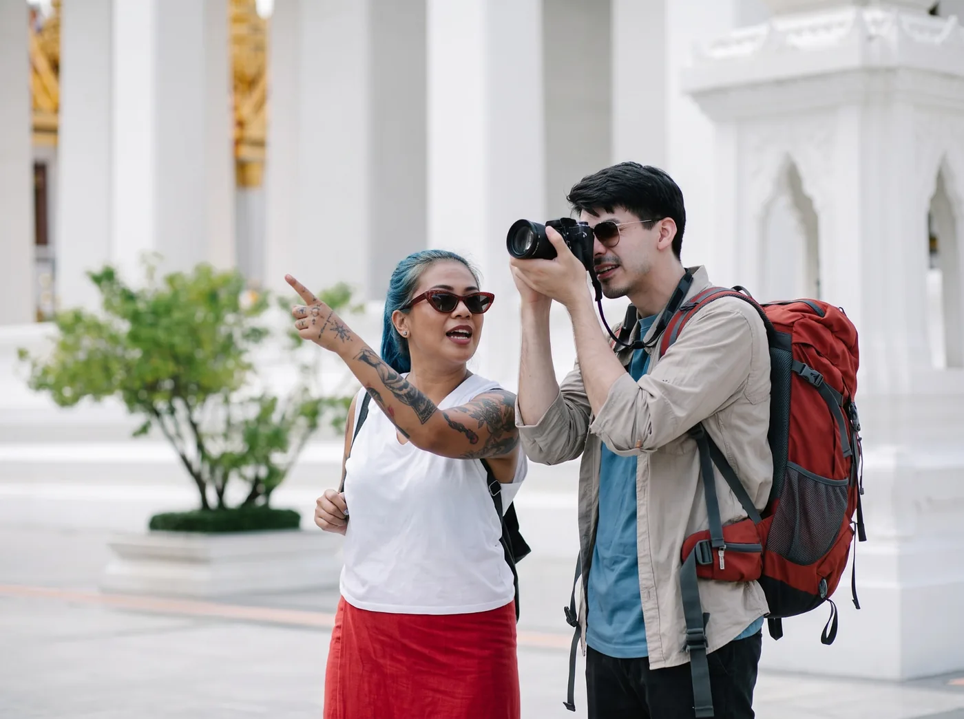 Dois turistas com mochilas fotografando um ponto turístico enquanto conversam em inglês